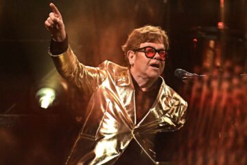Elton John no palco usando blazer dourado e óculos vermelhos, apontando para o alto enquanto canta ao piano.