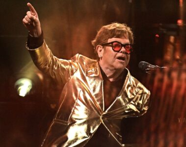 Elton John no palco usando blazer dourado e óculos vermelhos, apontando para o alto enquanto canta ao piano.