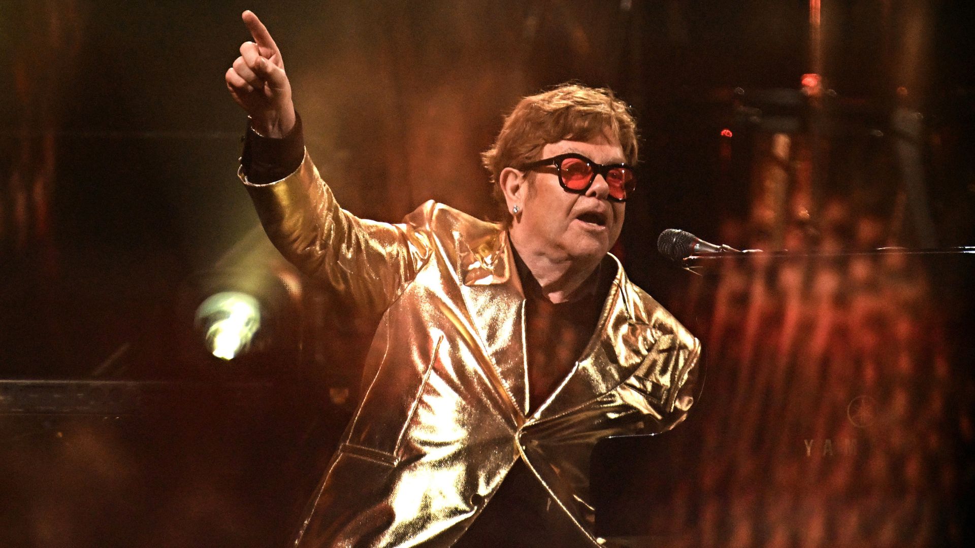 Elton John no palco usando blazer dourado e óculos vermelhos, apontando para o alto enquanto canta ao piano.