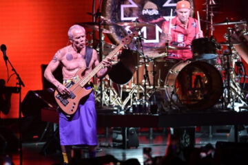 Flea tocando baixo no palco ao lado do baterista Chad Smith durante show do Red Hot Chili Peppers, com iluminação vermelha ao fundo.