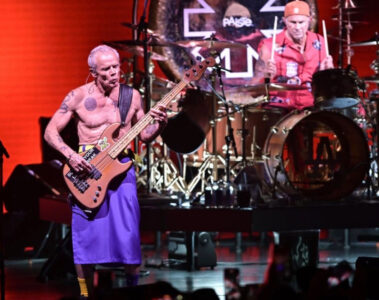 Flea tocando baixo no palco ao lado do baterista Chad Smith durante show do Red Hot Chili Peppers, com iluminação vermelha ao fundo.