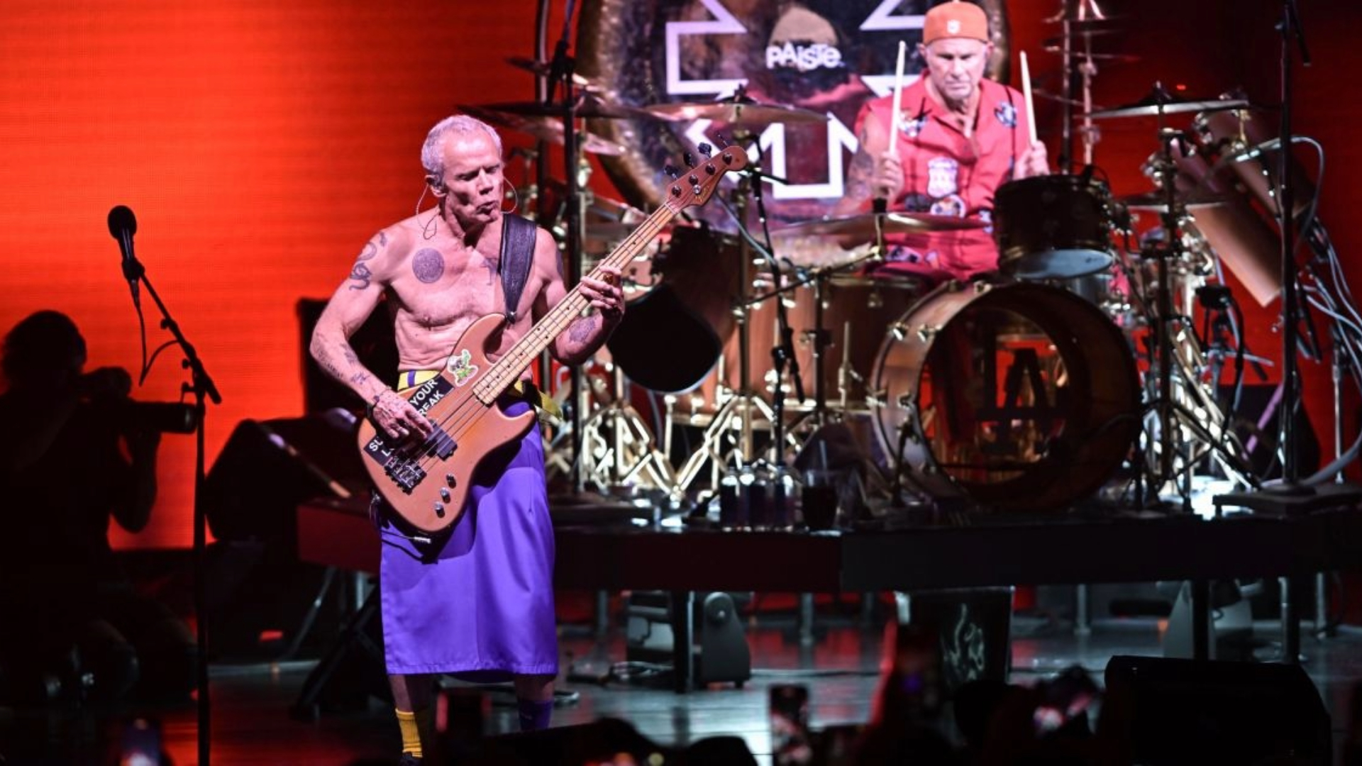 Flea tocando baixo no palco ao lado do baterista Chad Smith durante show do Red Hot Chili Peppers, com iluminação vermelha ao fundo.