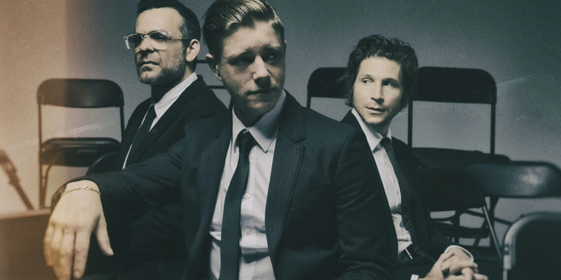 Interpol em ensaio fotográfico oficial, trio sentado em cadeiras, usando ternos escuros — divulgação do sideshow no Lollapalooza Brasil 2026.