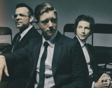 Interpol em ensaio fotográfico oficial, trio sentado em cadeiras, usando ternos escuros — divulgação do sideshow no Lollapalooza Brasil 2026.