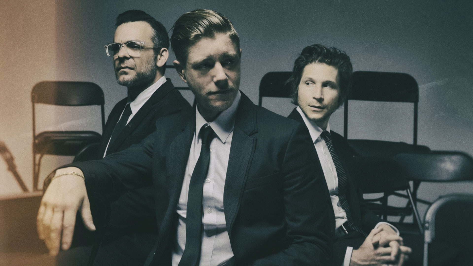 Interpol em ensaio fotográfico oficial, trio sentado em cadeiras, usando ternos escuros — divulgação do sideshow no Lollapalooza Brasil 2026.