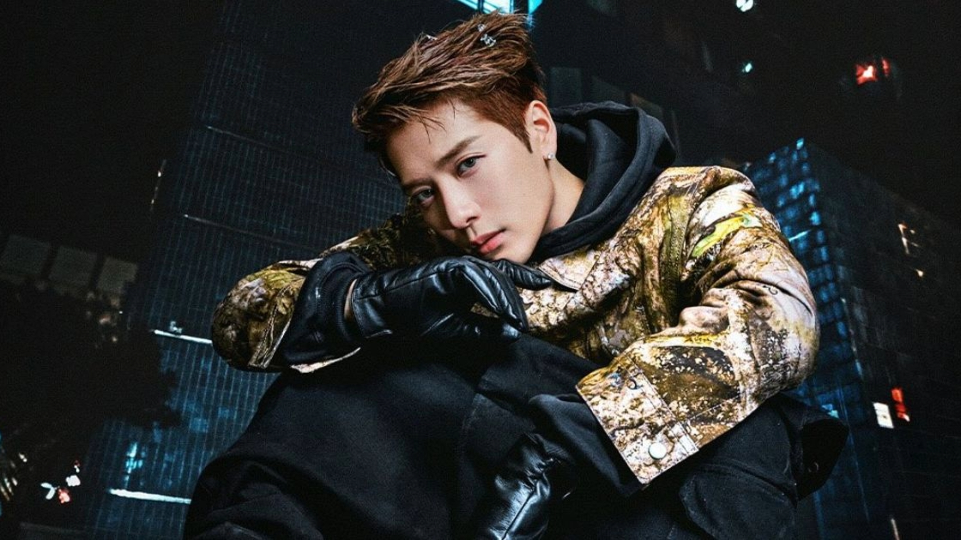 Jackson Wang posando com casaco estampado e luvas pretas em ambiente urbano à noite.