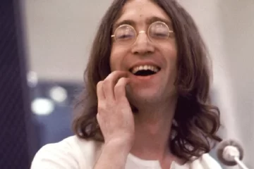John Lennon sorrindo durante gravação, usando óculos redondos e camisa branca, em uma foto de estúdio. Foto: © Apple Corps Ltd.