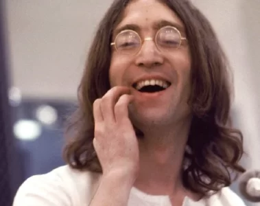 John Lennon sorrindo durante gravação, usando óculos redondos e camisa branca, em uma foto de estúdio. Foto: © Apple Corps Ltd.