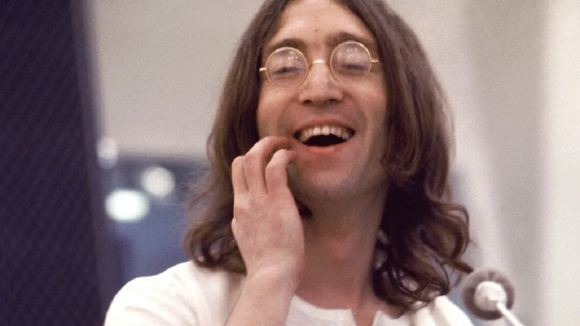 John Lennon sorrindo durante gravação, usando óculos redondos e camisa branca, em uma foto de estúdio. Foto: © Apple Corps Ltd.