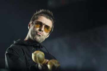 Liam Gallagher no palco usando óculos amarelos e segurando instrumentos de percussão.