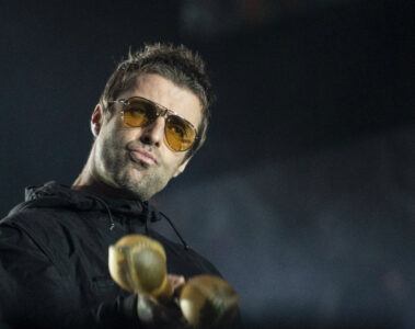 Liam Gallagher no palco usando óculos amarelos e segurando instrumentos de percussão.