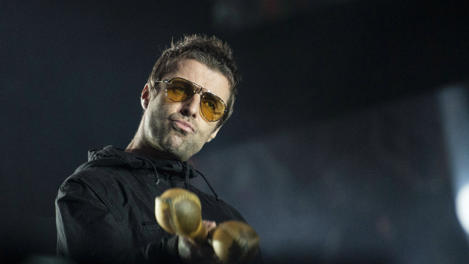 Liam Gallagher no palco usando óculos amarelos e segurando instrumentos de percussão.