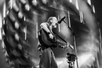 Thom Yorke tocando violão e cantando em um show do Radiohead, com iluminação intensa no palco. Foto: @twoshortdays.