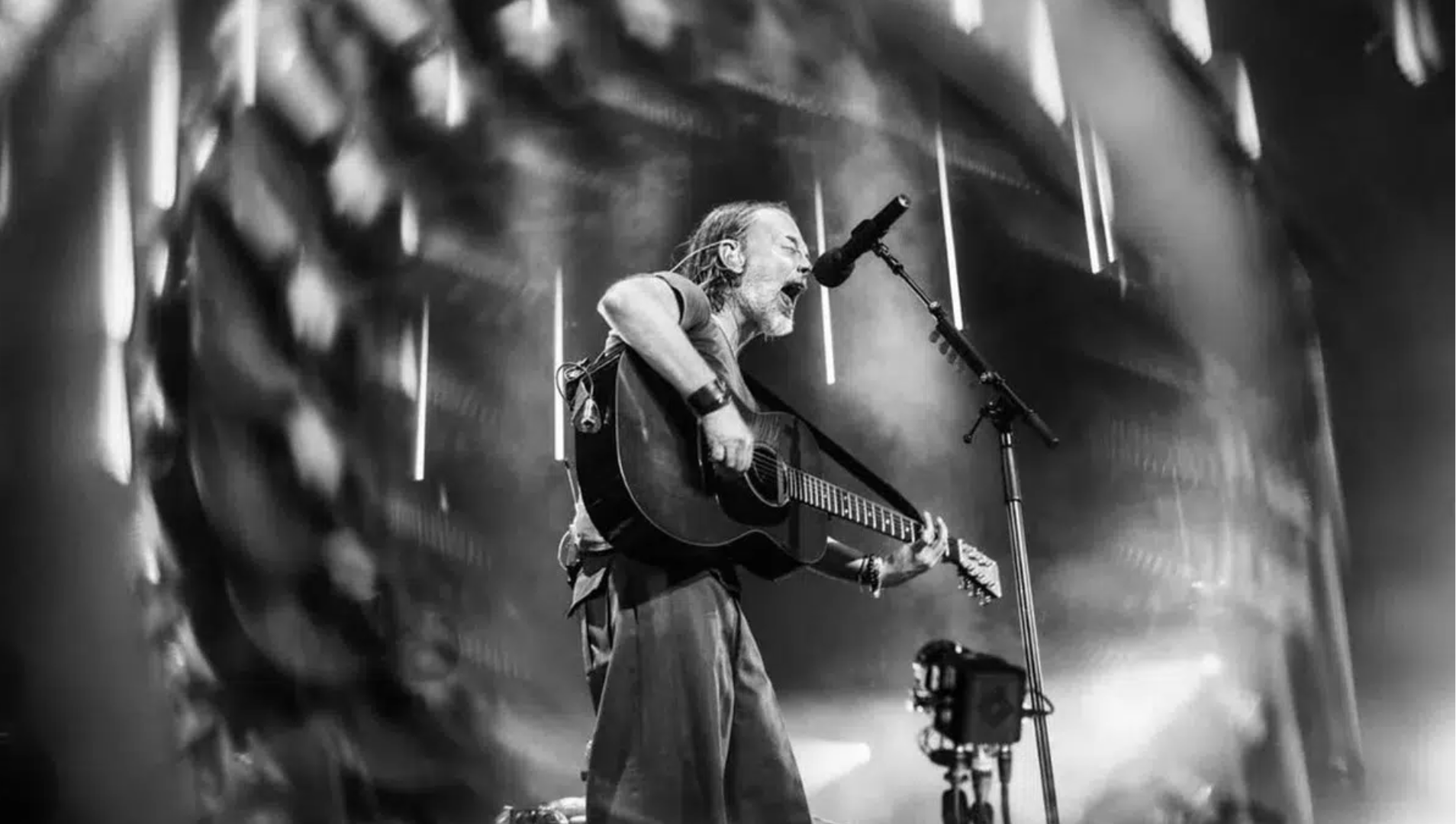 Thom Yorke tocando violão e cantando em um show do Radiohead, com iluminação intensa no palco. Foto: @twoshortdays.