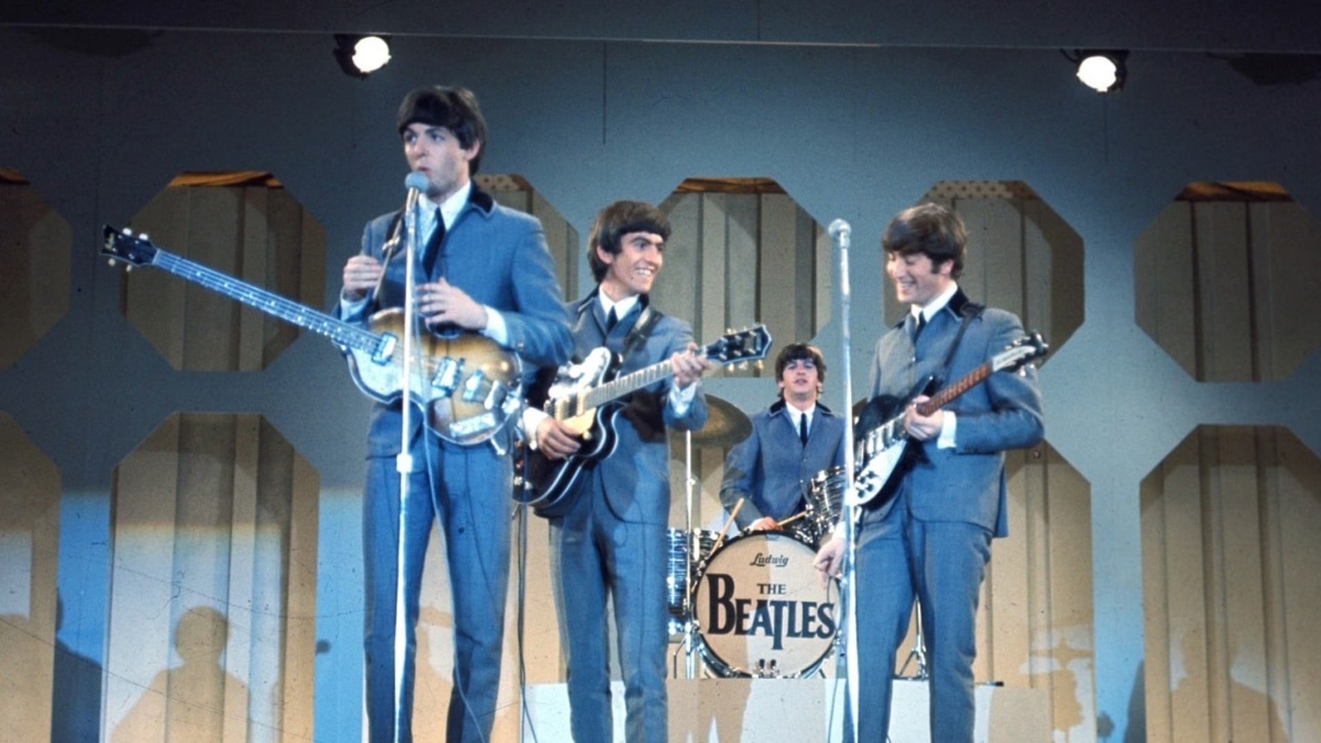 Os Beatles se apresentando ao vivo nos anos 60, com Paul McCartney, George Harrison, John Lennon e Ringo Starr no palco com seus instrumentos.