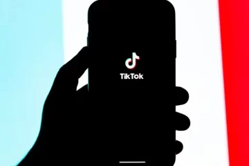Mão segurando um smartphone com o logo do TikTok na tela, em frente a um fundo colorido em azul e vermelho.