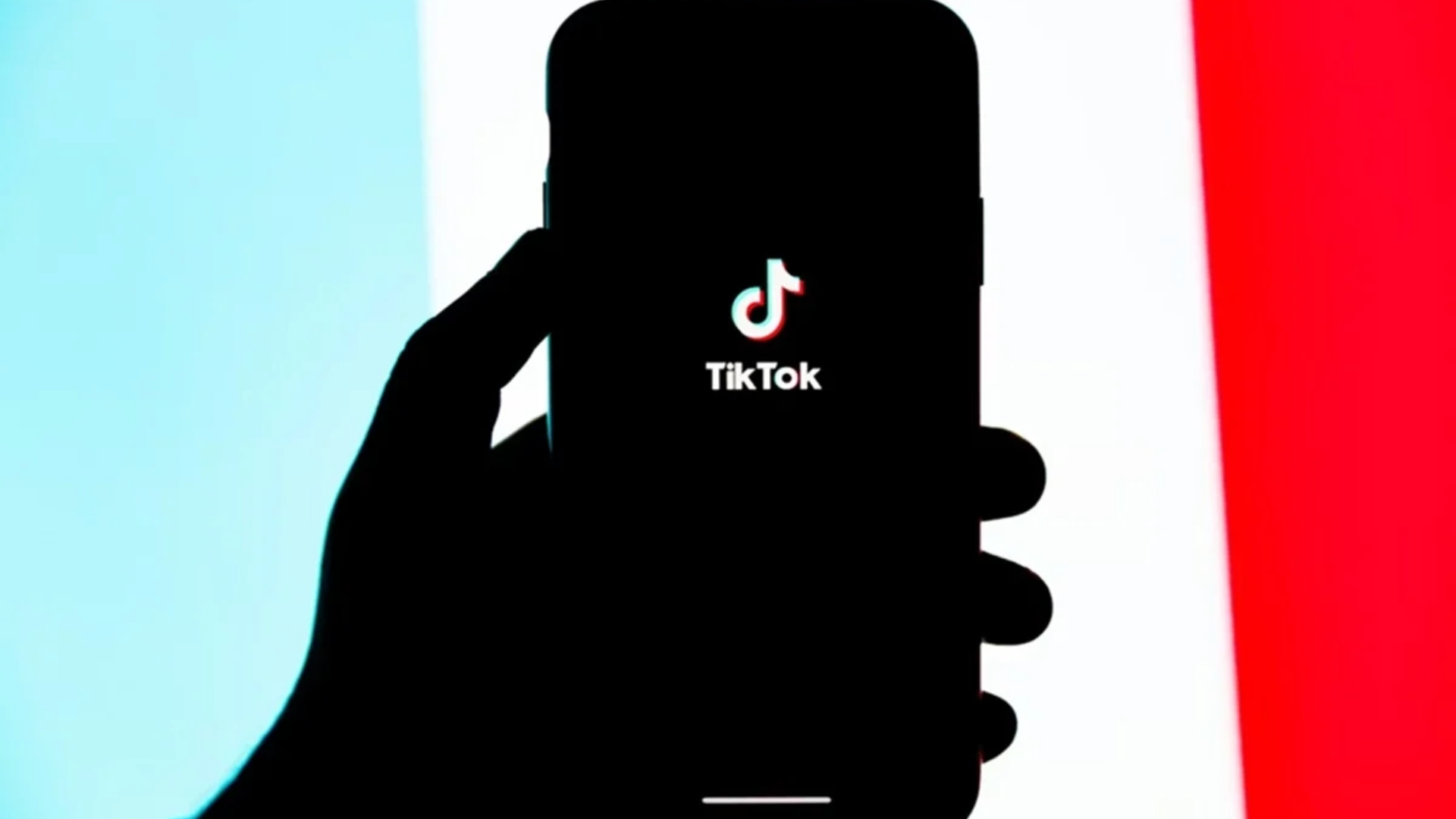 Mão segurando um smartphone com o logo do TikTok na tela, em frente a um fundo colorido em azul e vermelho.