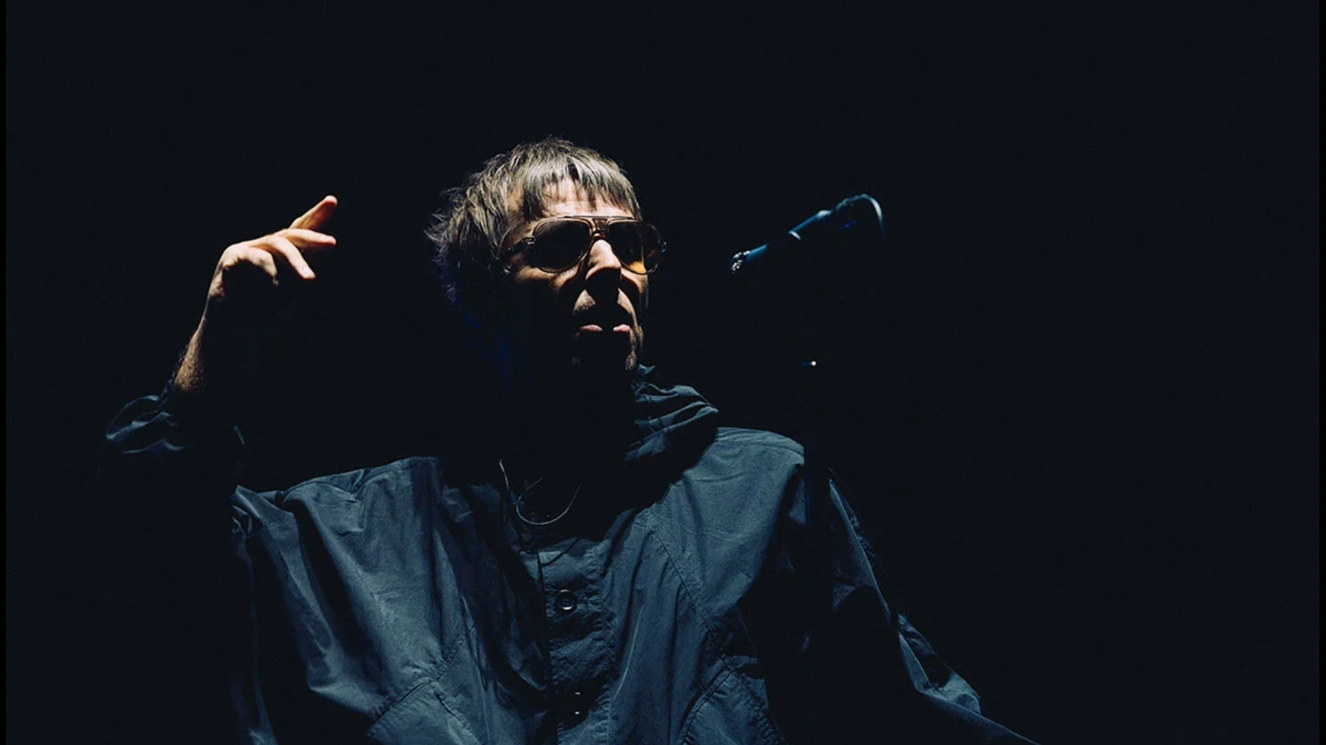 Liam Gallagher durante apresentação ao vivo do Oasis, sob iluminação baixa no palco.
