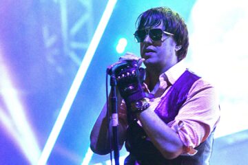 Julian Casablancas cantando ao vivo com o The Strokes, usando óculos escuros e segurando o microfone sob luzes coloridas de palco.