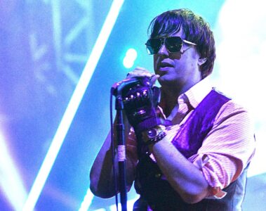 Julian Casablancas cantando ao vivo com o The Strokes, usando óculos escuros e segurando o microfone sob luzes coloridas de palco.