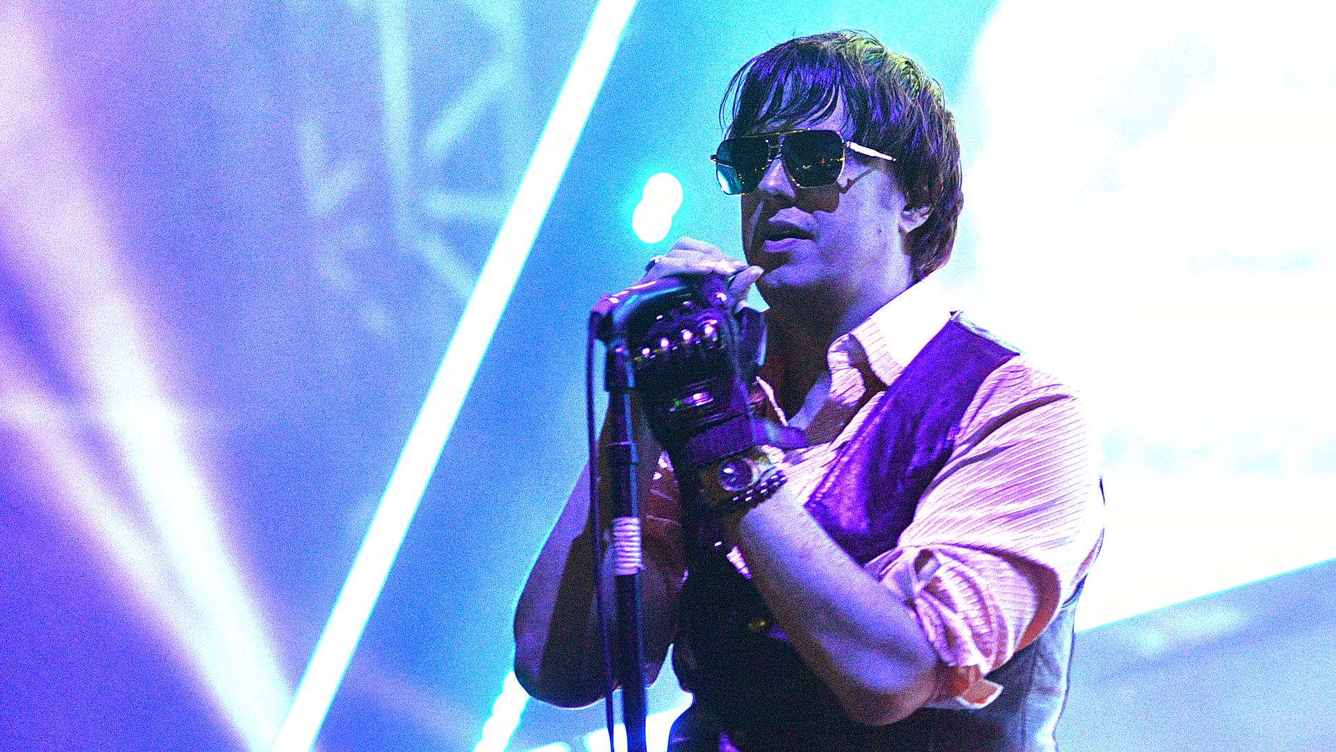 Julian Casablancas cantando ao vivo com o The Strokes, usando óculos escuros e segurando o microfone sob luzes coloridas de palco.