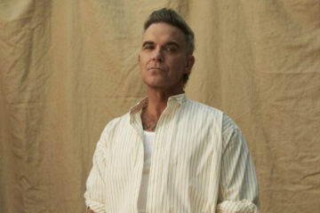 Robbie Williams em ensaio promocional, vestindo camisa clara e posando diante de fundo neutro.