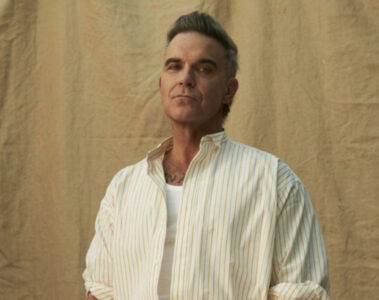 Robbie Williams em ensaio promocional, vestindo camisa clara e posando diante de fundo neutro.