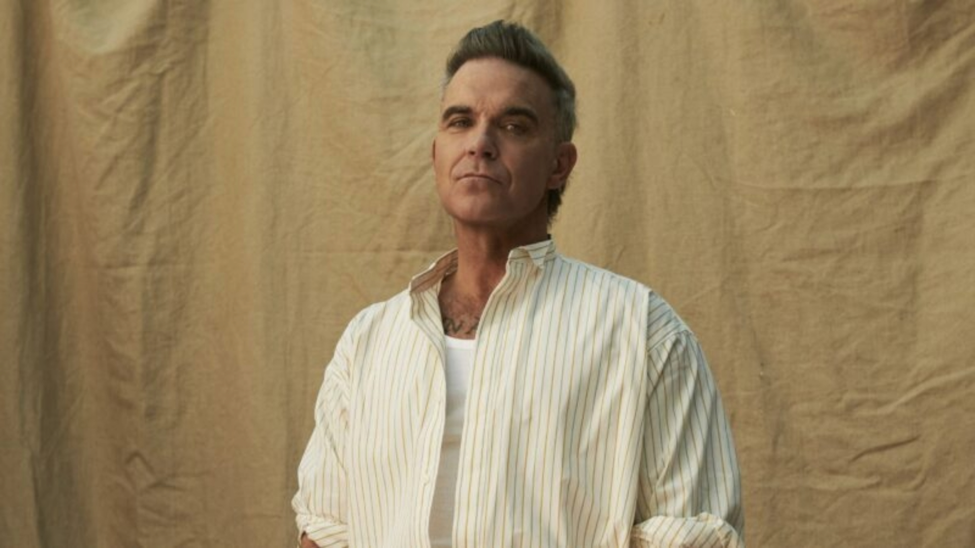 Robbie Williams em ensaio promocional, vestindo camisa clara e posando diante de fundo neutro.