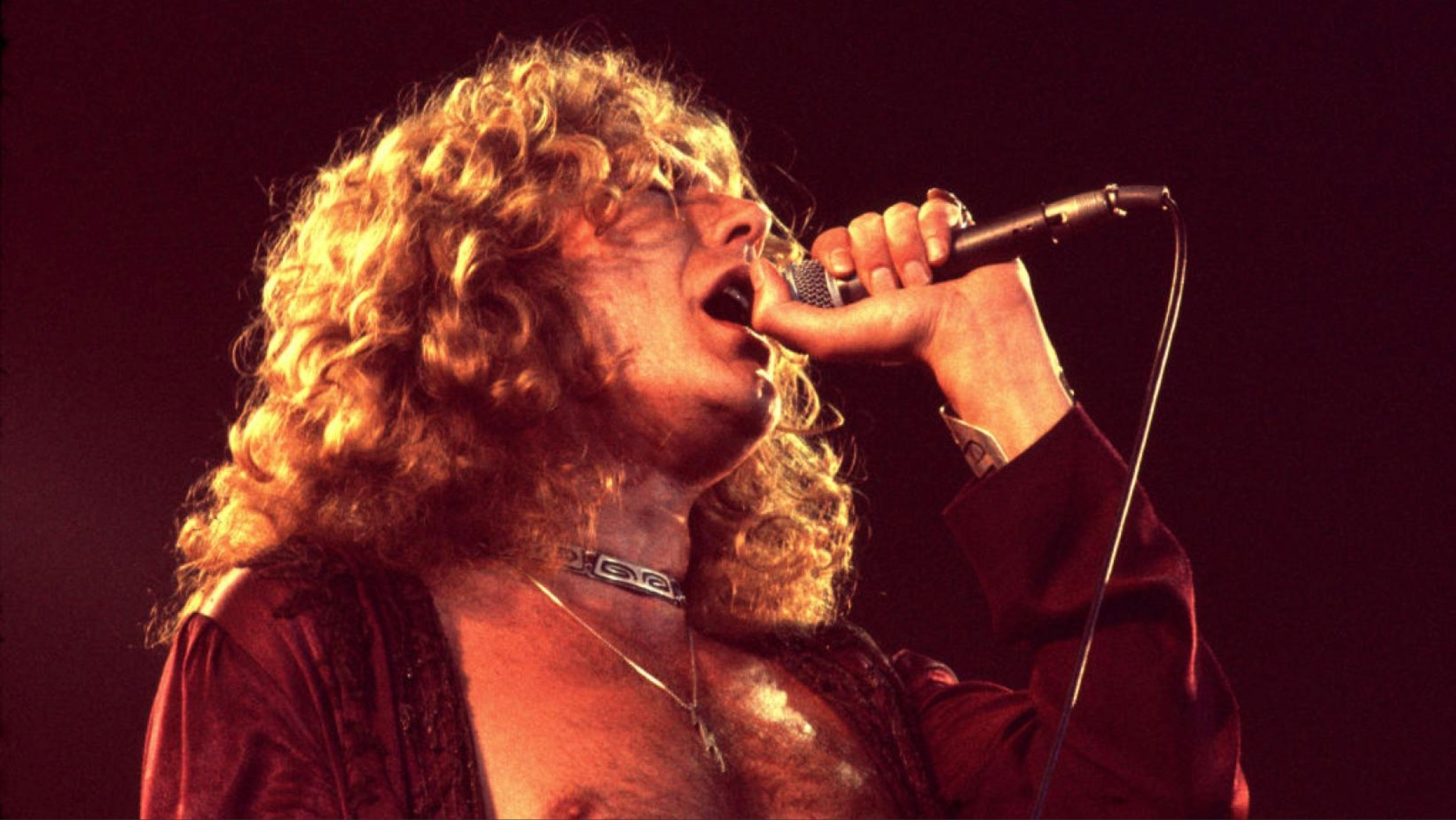 Robert Plant cantando ao vivo nos anos 70, durante apresentação do Led Zeppelin, segurando o microfone com intensidade.