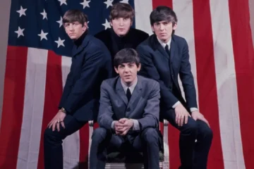 Os Beatles posam em frente à bandeira dos Estados Unidos durante a fase inicial da Beatlemania em 1964