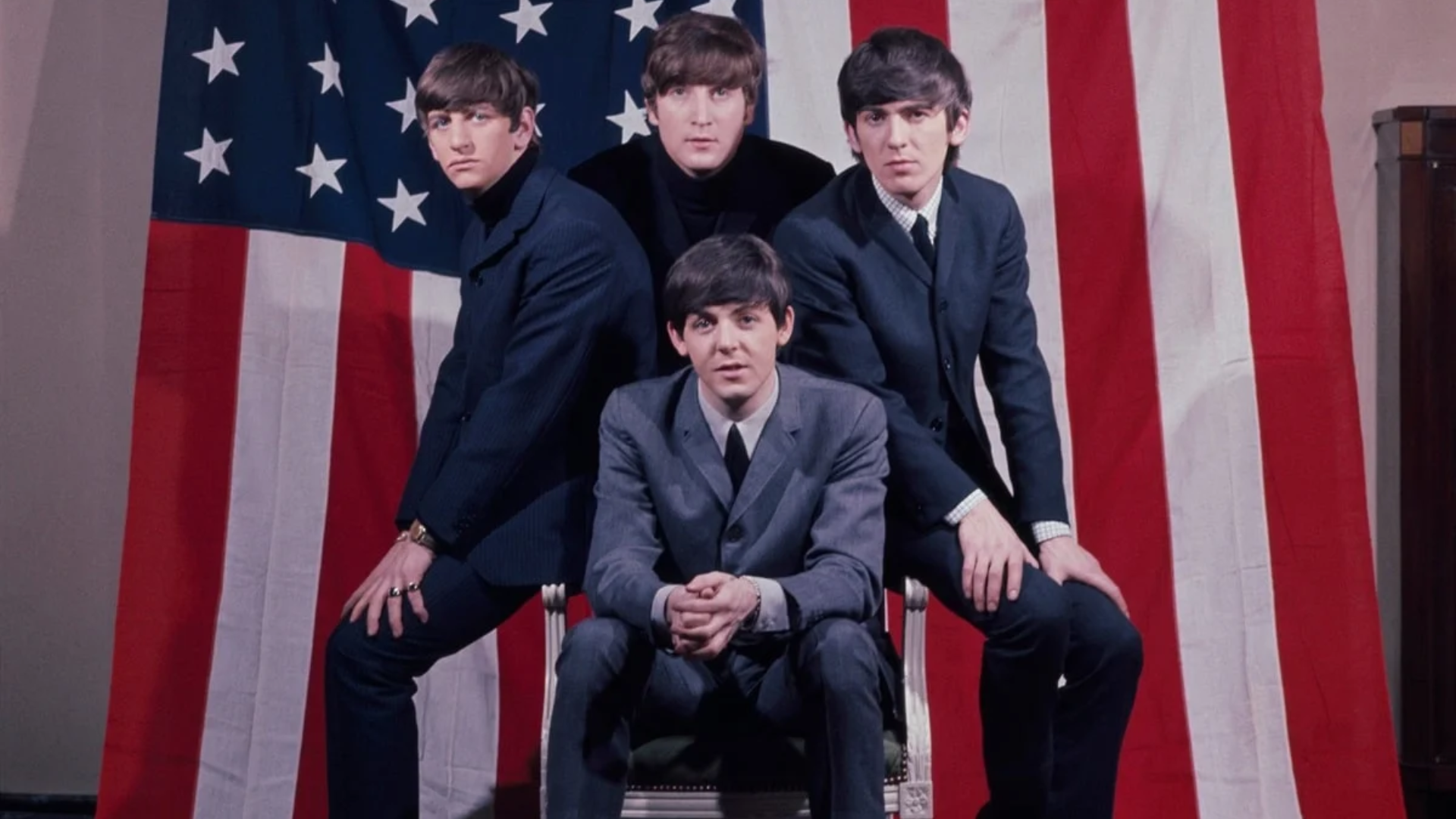 Os Beatles posam em frente à bandeira dos Estados Unidos durante a fase inicial da Beatlemania em 1964