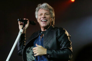 Jon Bon Jovi sorri enquanto canta em um palco iluminado, segurando um microfone durante uma apresentação ao vivo.