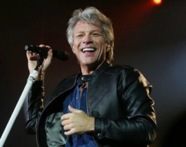 Jon Bon Jovi sorri enquanto canta em um palco iluminado, segurando um microfone durante uma apresentação ao vivo.