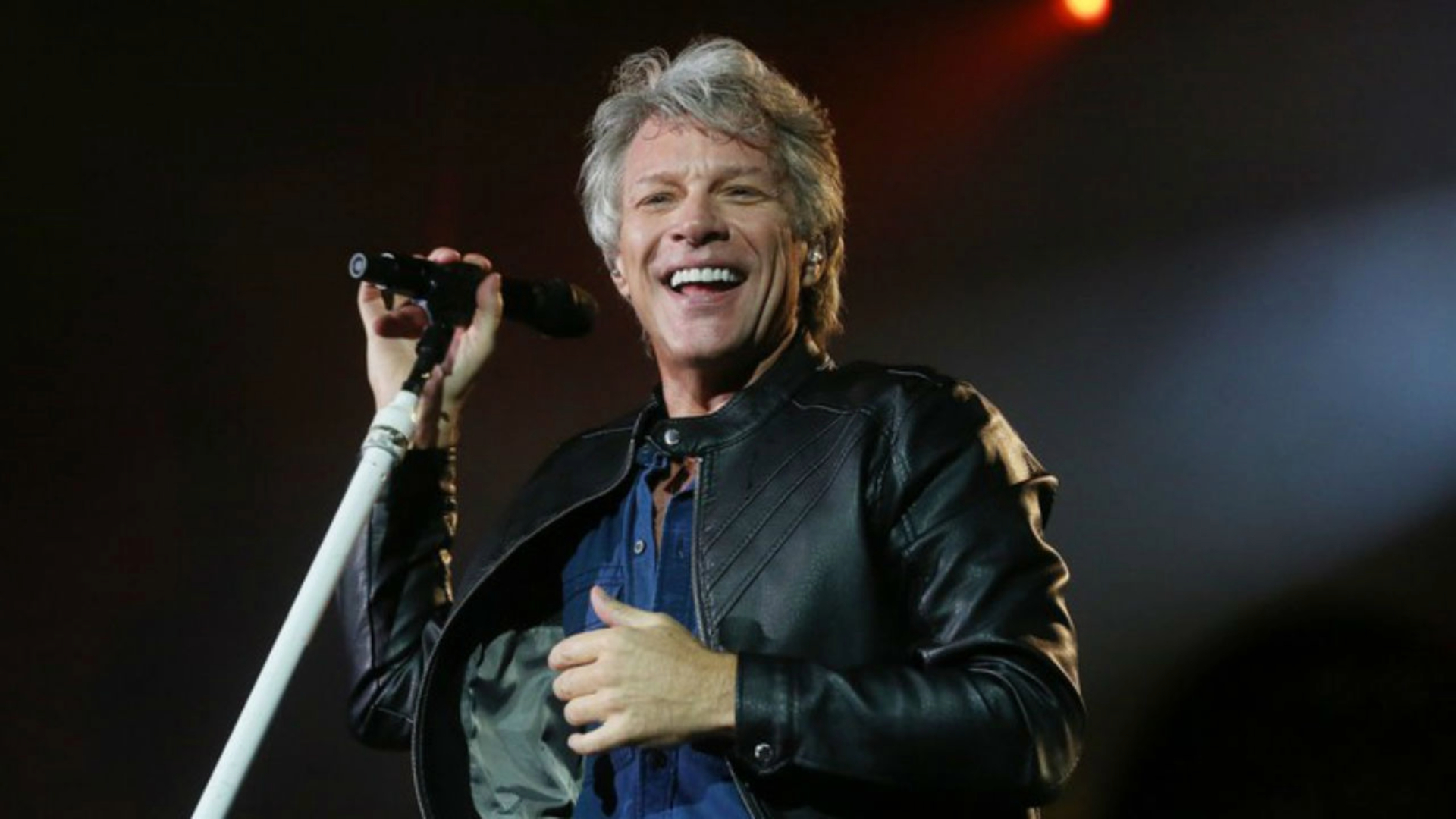 Jon Bon Jovi sorri enquanto canta em um palco iluminado, segurando um microfone durante uma apresentação ao vivo.