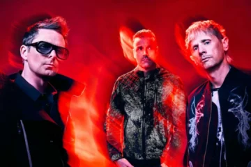Integrantes do Muse posam diante de um fundo vermelho com efeitos de luz durante a divulgação do single Nightshift Superstar e do álbum The Wow! Signal.