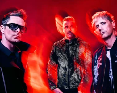Integrantes do Muse posam diante de um fundo vermelho com efeitos de luz durante a divulgação do single Nightshift Superstar e do álbum The Wow! Signal.