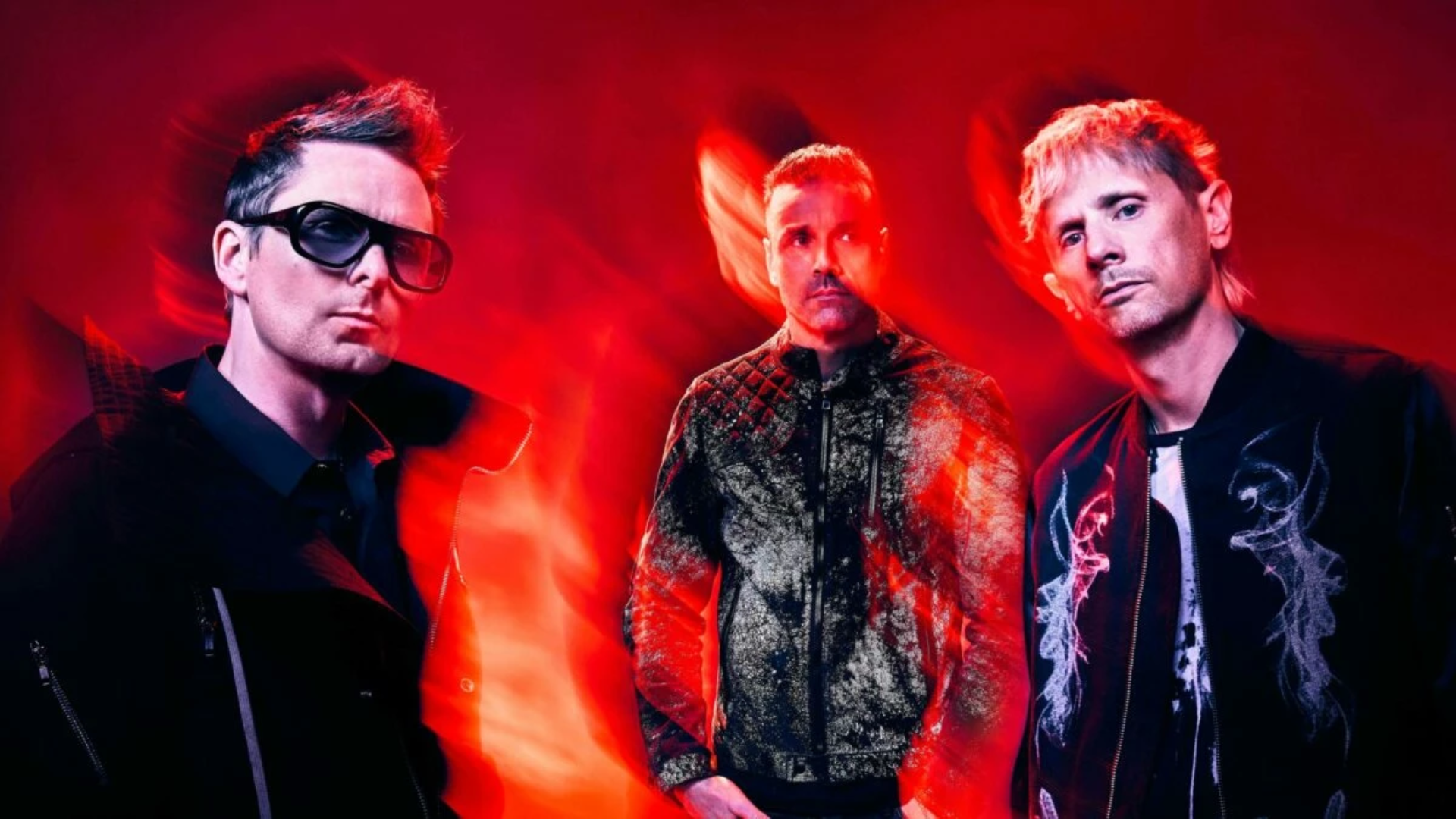 Integrantes do Muse posam diante de um fundo vermelho com efeitos de luz durante a divulgação do single Nightshift Superstar e do álbum The Wow! Signal.