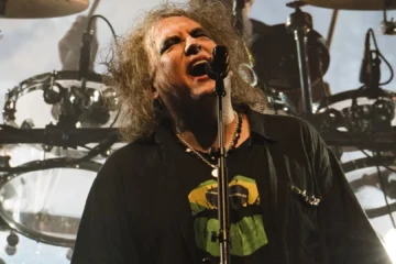 Robert Smith se apresenta ao vivo com o The Cure durante show da banda, em meio a uma iluminação intensa e bateria ao fundo.