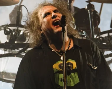 Robert Smith se apresenta ao vivo com o The Cure durante show da banda, em meio a uma iluminação intensa e bateria ao fundo.