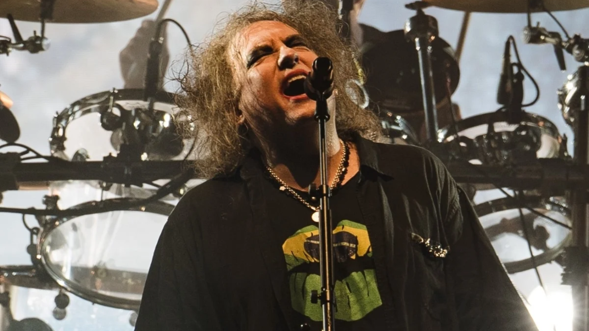 Robert Smith se apresenta ao vivo com o The Cure durante show da banda, em meio a uma iluminação intensa e bateria ao fundo.