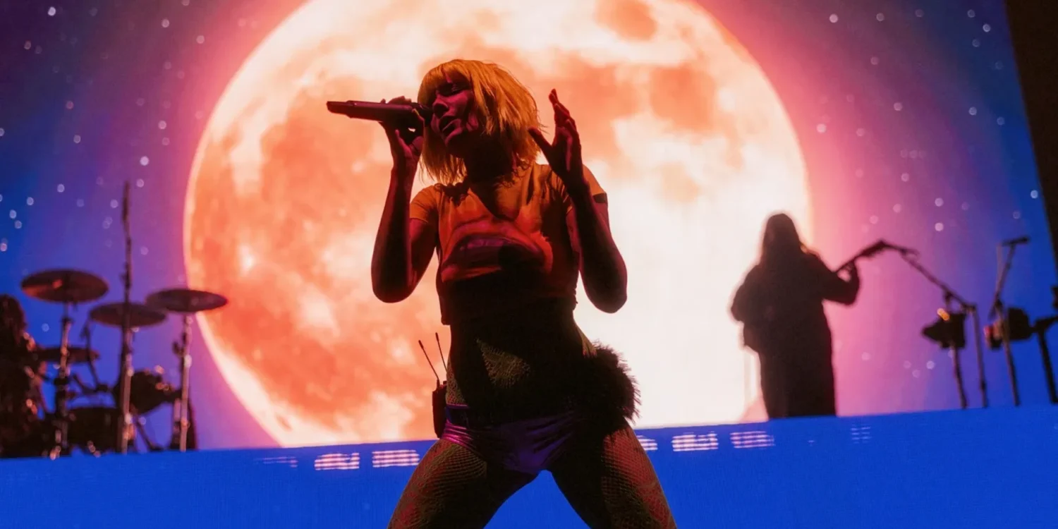 Halsey canta no palco durante uma apresentação com uma grande lua iluminada ao fundo.
