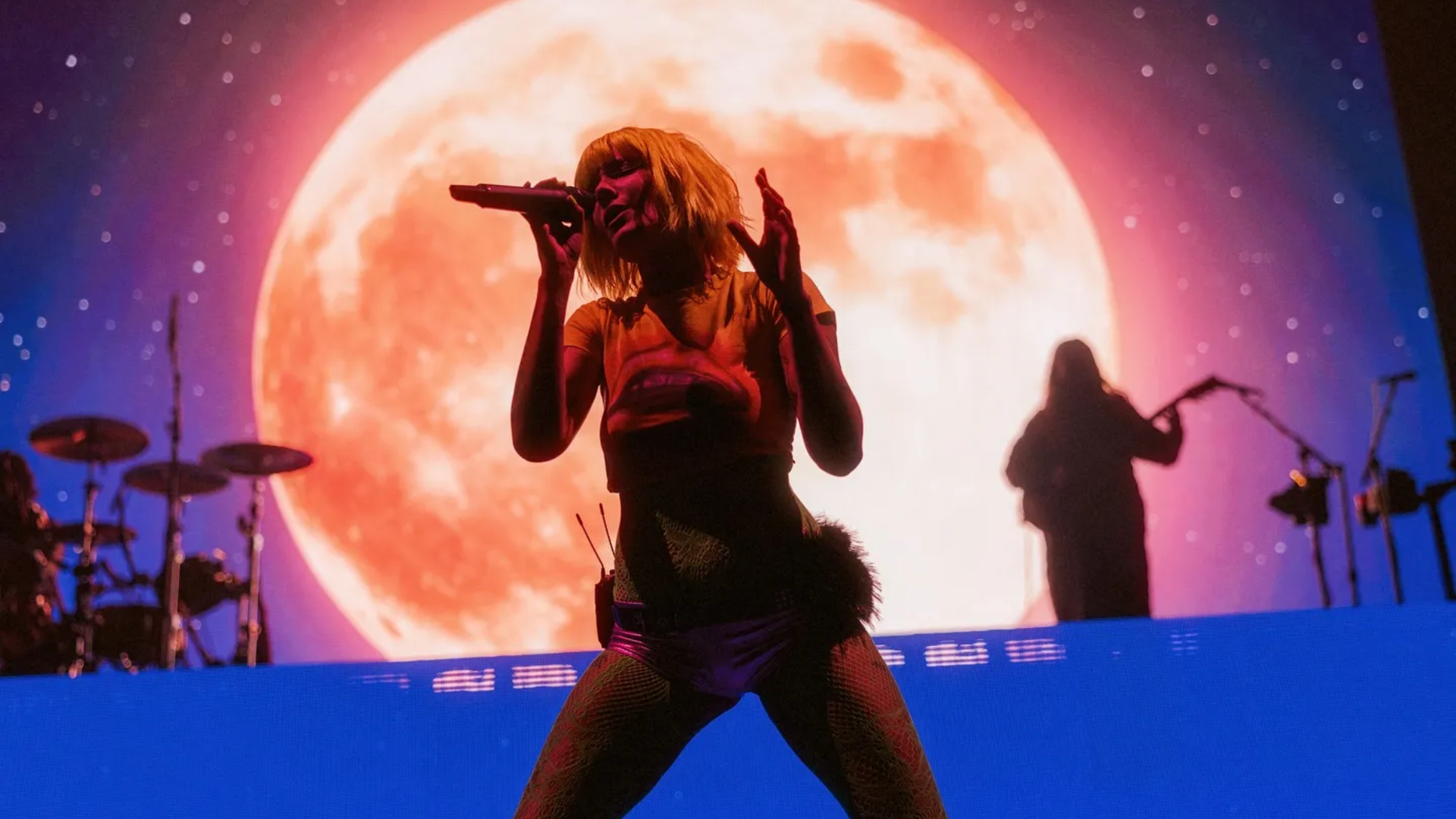 Halsey canta no palco durante uma apresentação com uma grande lua iluminada ao fundo.
