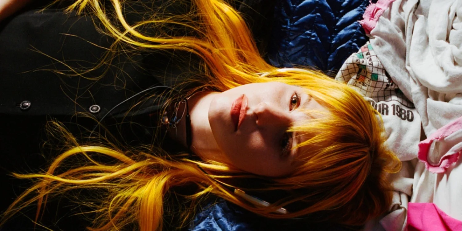 Hayley Williams aparece deitada, com cabelos longos alaranjados espalhados sobre tecidos azuis, brancos e rosas, olhando para a câmera.