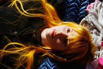 Hayley Williams aparece deitada, com cabelos longos alaranjados espalhados sobre tecidos azuis, brancos e rosas, olhando para a câmera.