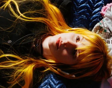 Hayley Williams aparece deitada, com cabelos longos alaranjados espalhados sobre tecidos azuis, brancos e rosas, olhando para a câmera.
