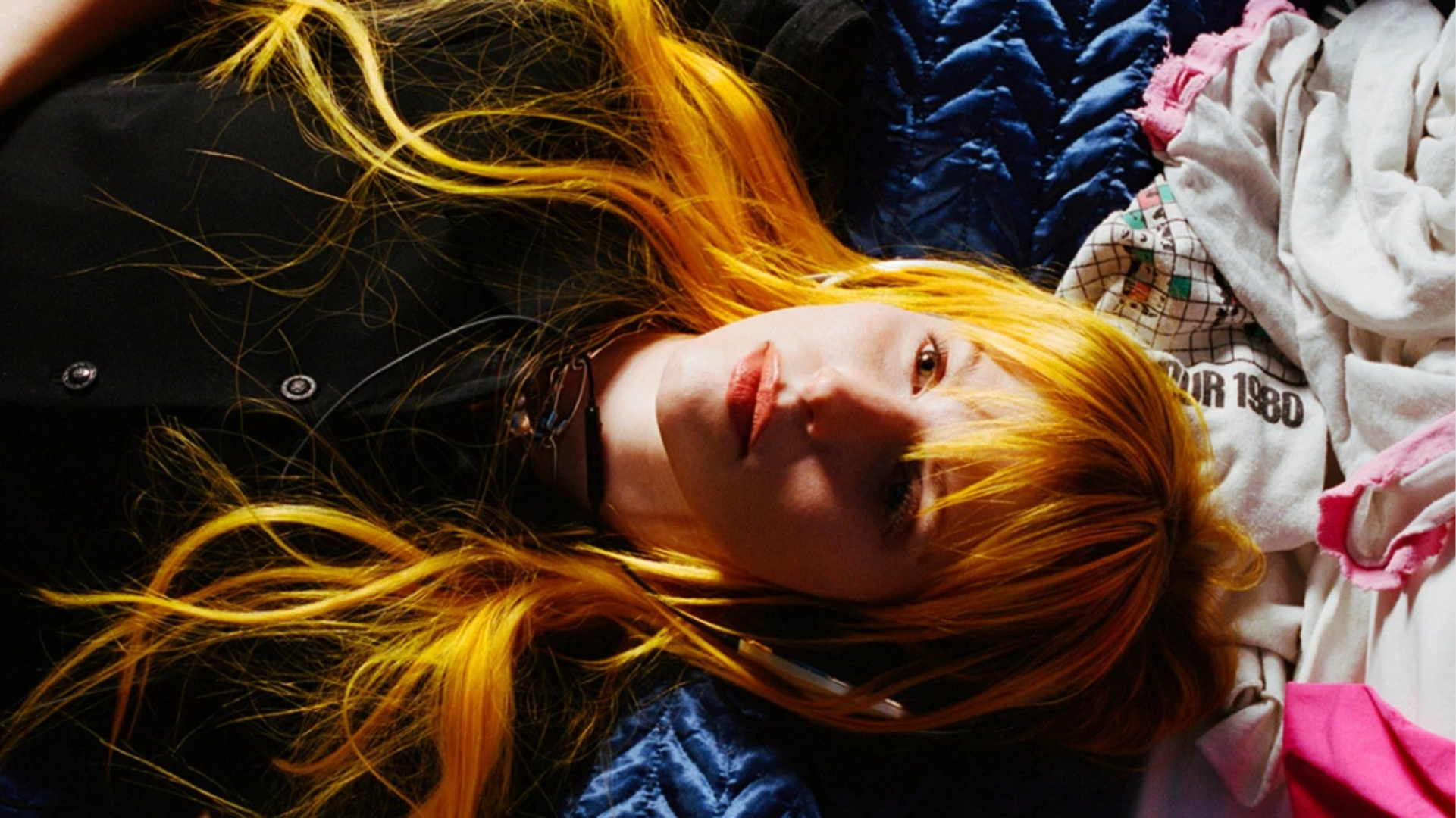 Hayley Williams aparece deitada, com cabelos longos alaranjados espalhados sobre tecidos azuis, brancos e rosas, olhando para a câmera.