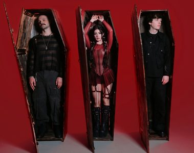Integrantes do Against The Current, com Chrissy Costanza ao centro, posam dentro de caixões de madeira diante de um fundo vermelho.