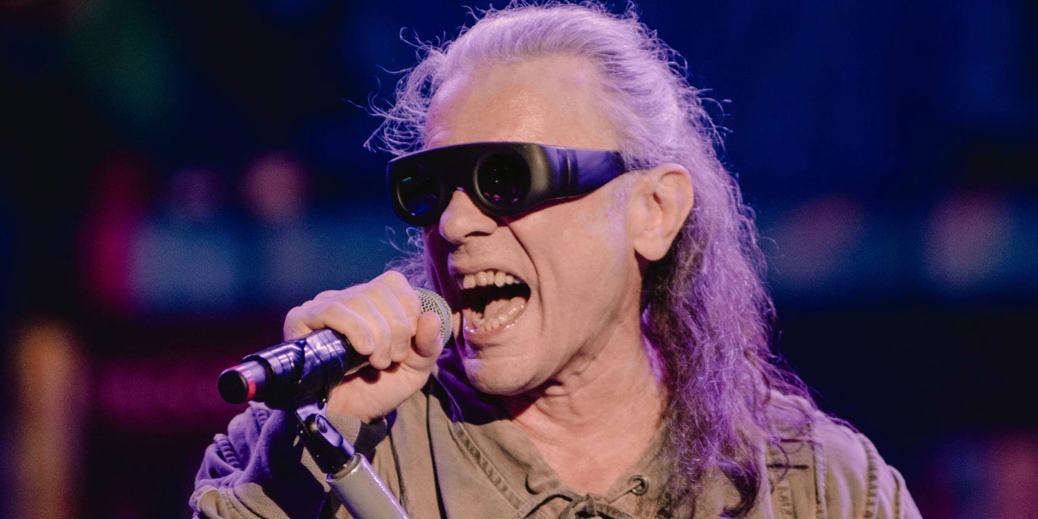 Bruce Dickinson, vocalista do Iron Maiden, cantando ao microfone durante apresentação ao vivo, usando óculos escuros e sob iluminação azul e roxa.