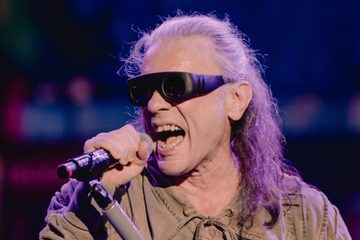 Bruce Dickinson, vocalista do Iron Maiden, cantando ao microfone durante apresentação ao vivo, usando óculos escuros e sob iluminação azul e roxa.
