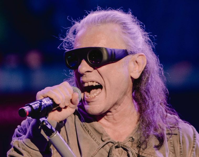 Bruce Dickinson, vocalista do Iron Maiden, cantando ao microfone durante apresentação ao vivo, usando óculos escuros e sob iluminação azul e roxa.